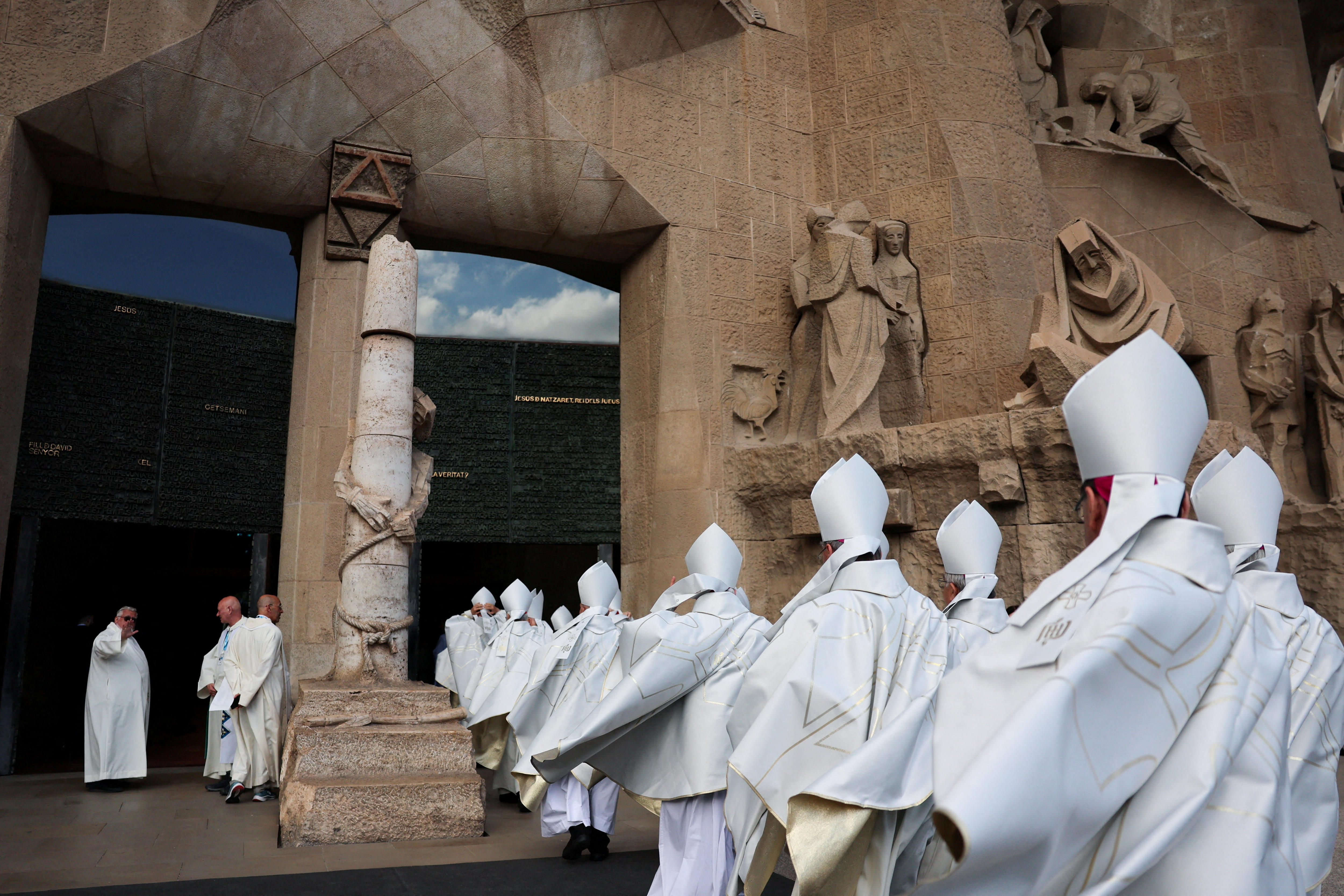 Catholic clergy members in white robes entering through a stone doorway surrounded by sculptures.