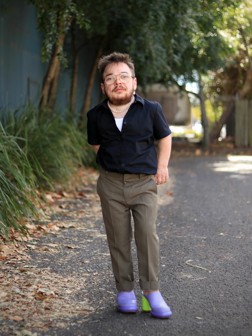A man, who has dwarfism, poses for the camera. He is wearing brightly-coloured neon heels.
