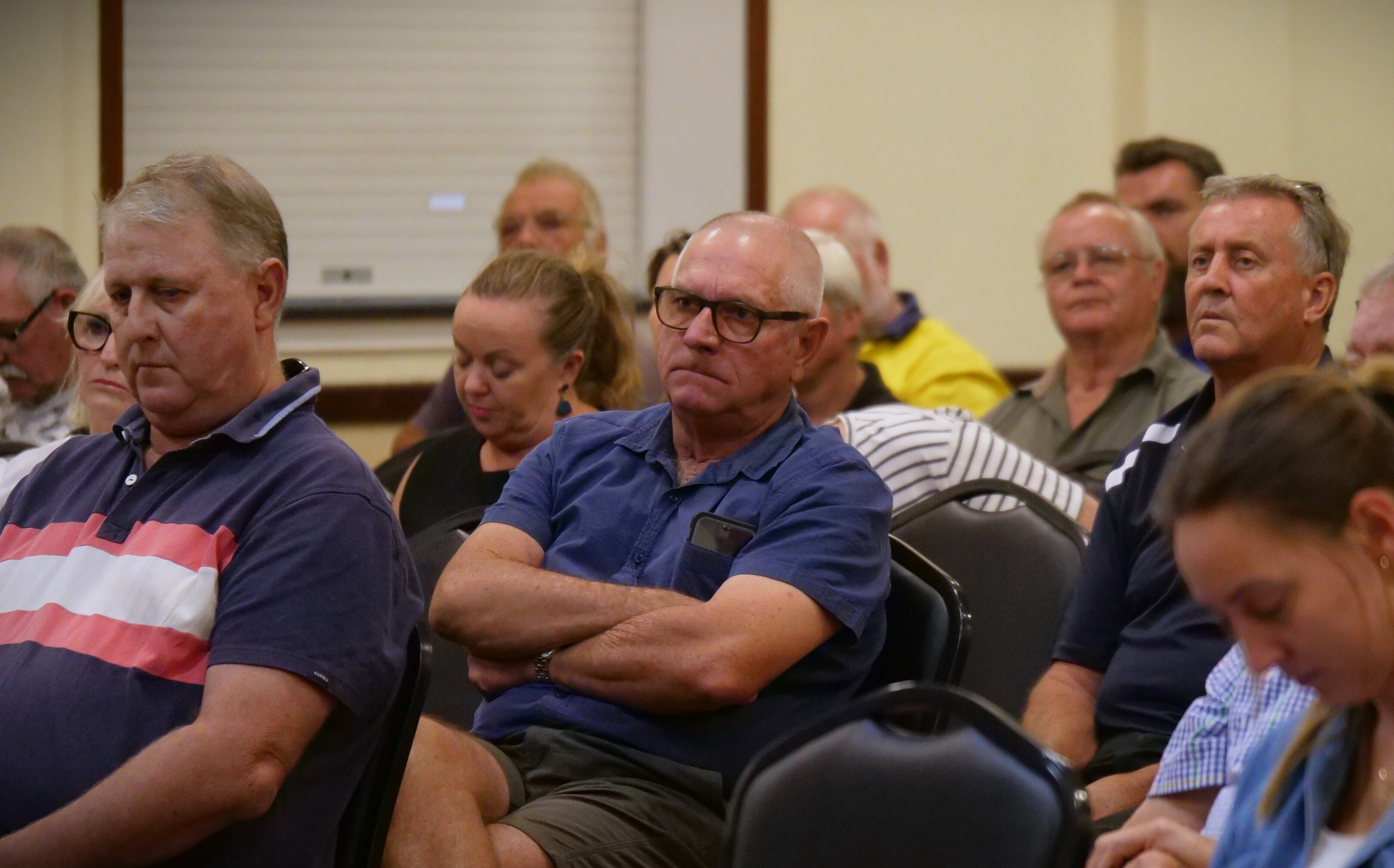 People sit on chairs inside. A man in the center wears a blue shirt and glasses with his arms crossed and a frown on his face.