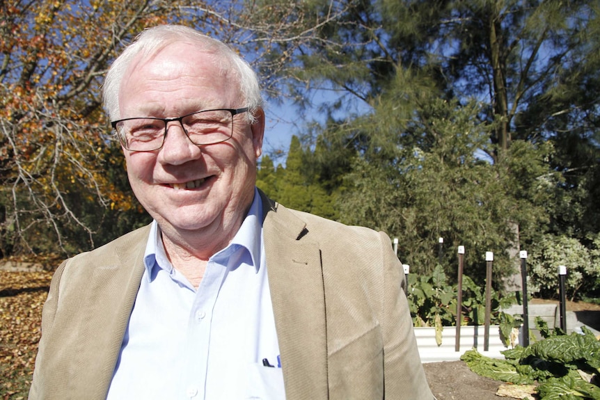 A man wearing a tan suit jacket smiles for a photo in a leafy garden.