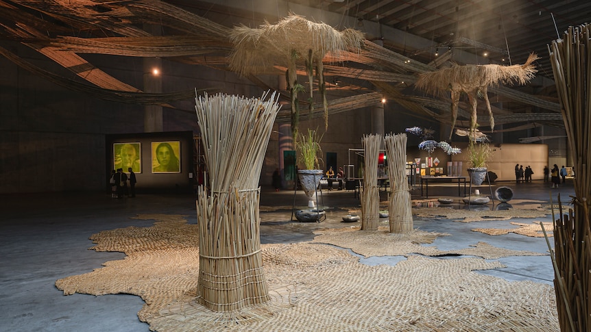 Reeds woven into mats are on the floor of a cavernous space, some reeds are bundled together and placed upright.