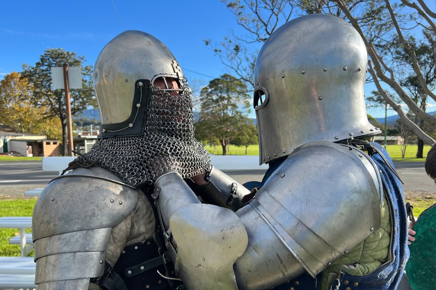 One man in full silver knight's armour helps another man in full armour secure his helmet which has chain mail over the mouth. 