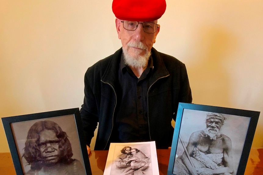 A man with a red beret hat is sitting at a table holding photos of his aboriginal ancestors and a map of his country.