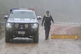 A policeman in a beanie stands next to a police car and tape and a road closed sign amid in a mountainous area.