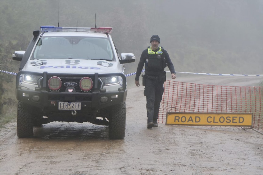 A policeman in a beanie stands next to a police car and tape and a road closed sign amid in a mountainous area.