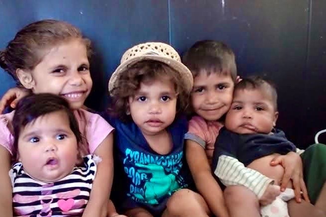 Five smiling young Aboriginal children hug each other tightly on a couch while looking directly at the camera.