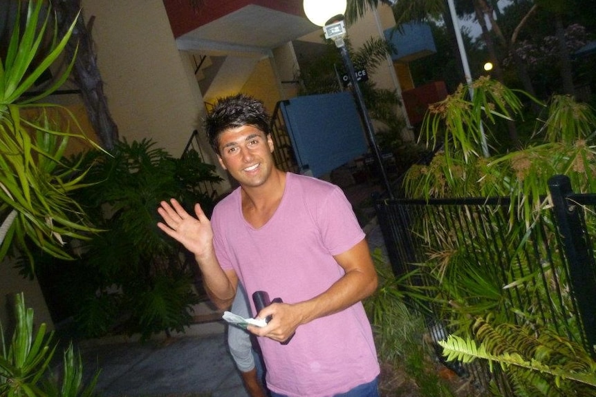 A young man in a pink t-shirt poses and waves toward the camera