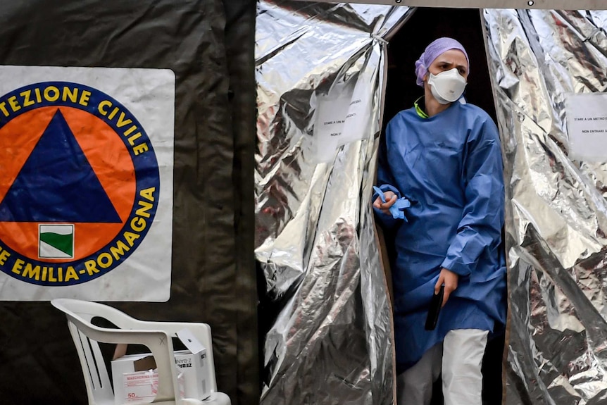 A woman wearing hospital scrubs and a face mask walks through the opening of a silvery tent.