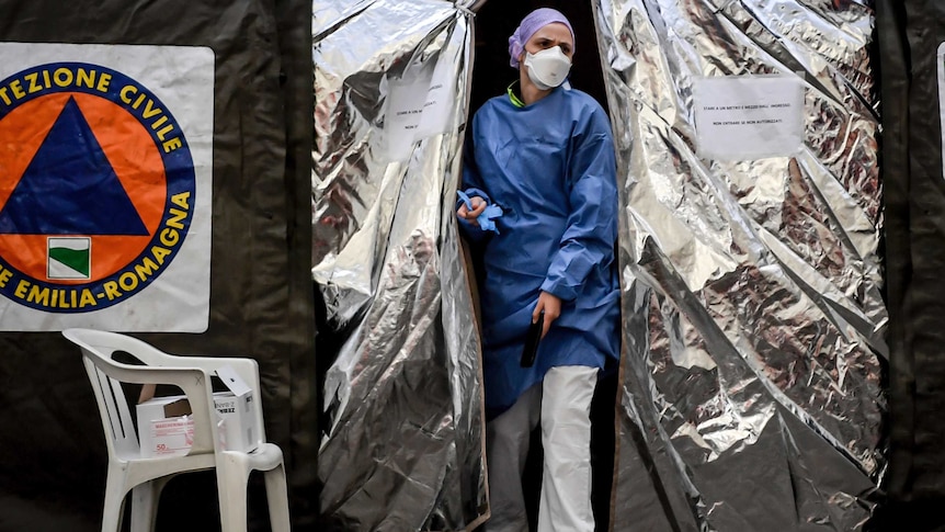 A woman wearing hospital scrubs and a face mask walks through the opening of a silvery tent.