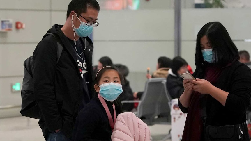 A family including a little girl wearing masks wait with their luggage as two men wearing masks also walk by with their luggage.