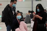 A family including a little girl wearing masks wait with their luggage as two men wearing masks also walk by with their luggage.