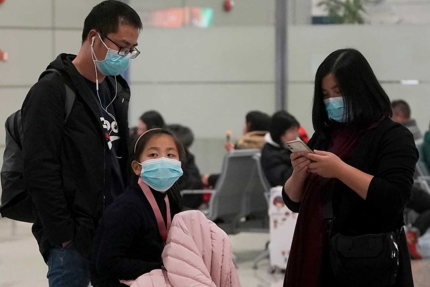 A family including a little girl wearing masks wait with their luggage as two men wearing masks also walk by with their luggage.