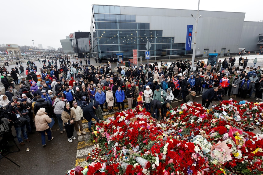 A large crowd surrounds a large collection of flowers, most red, placed on the ground.