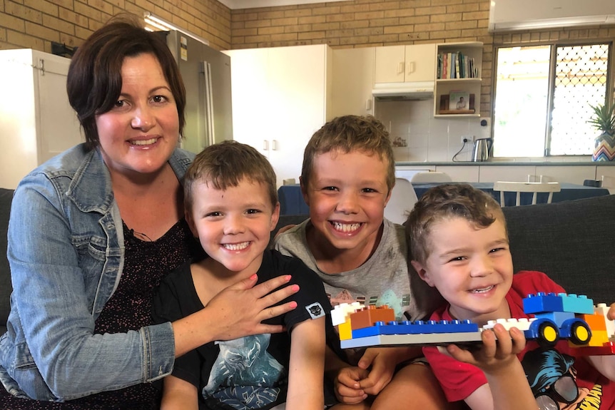 Stacey Hughes sits next to her three smiling boys on the couch in their home. One boy is holding a truck built from Lego pieces.