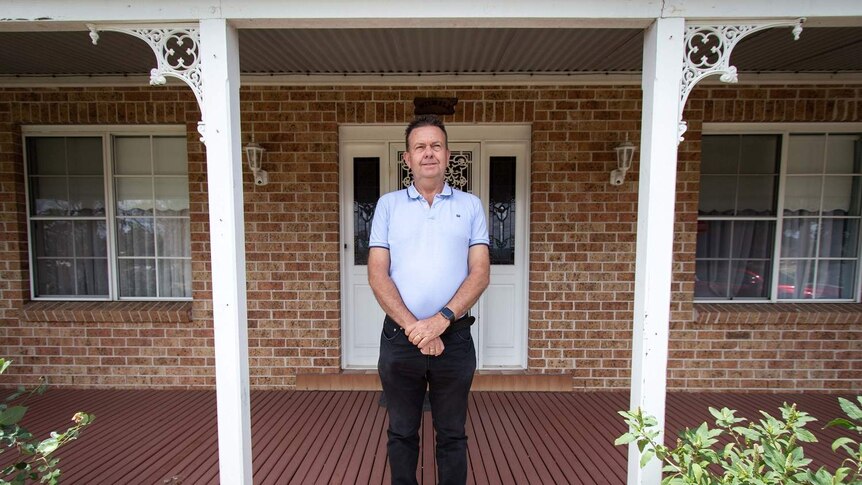 A man wearing a pale polo shirt and dark pants stands on a front porch with his hands crossed in front of him.
