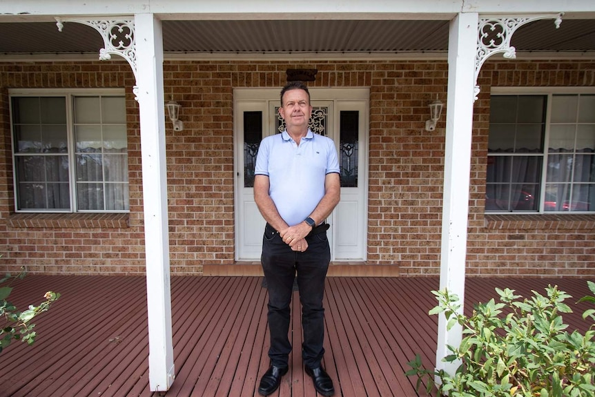 A man wearing a pale polo shirt and dark pants stands on a front porch with his hands crossed in front of him.