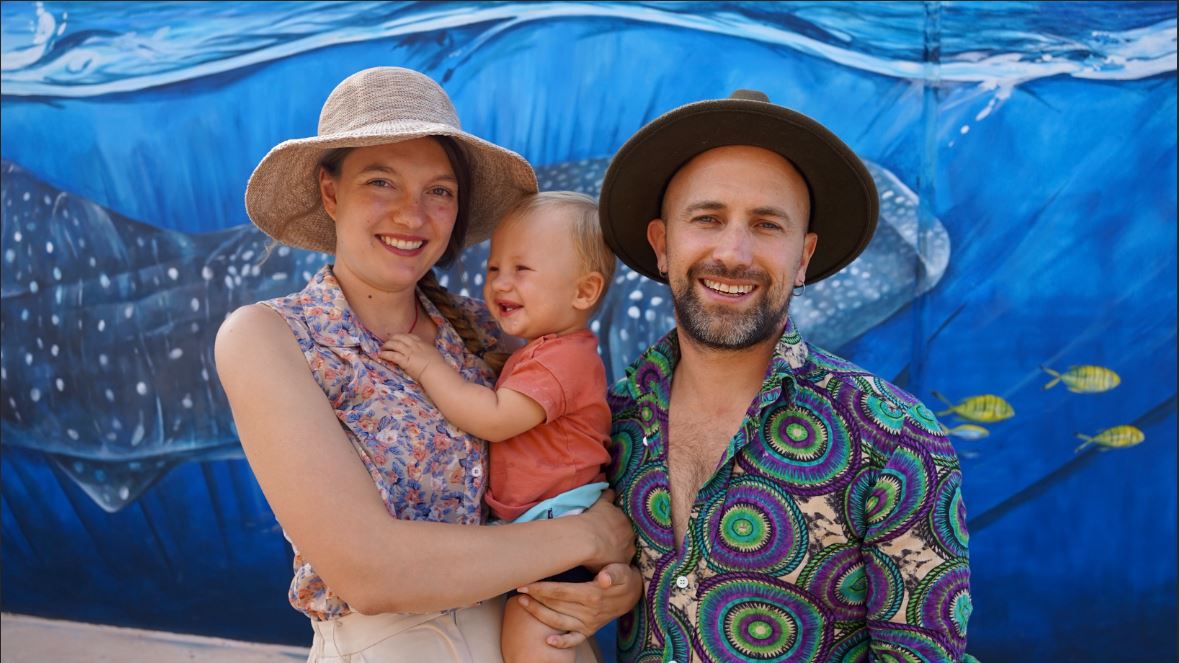 A man and woman wearing hats and colourful shirts holding a baby between them.
