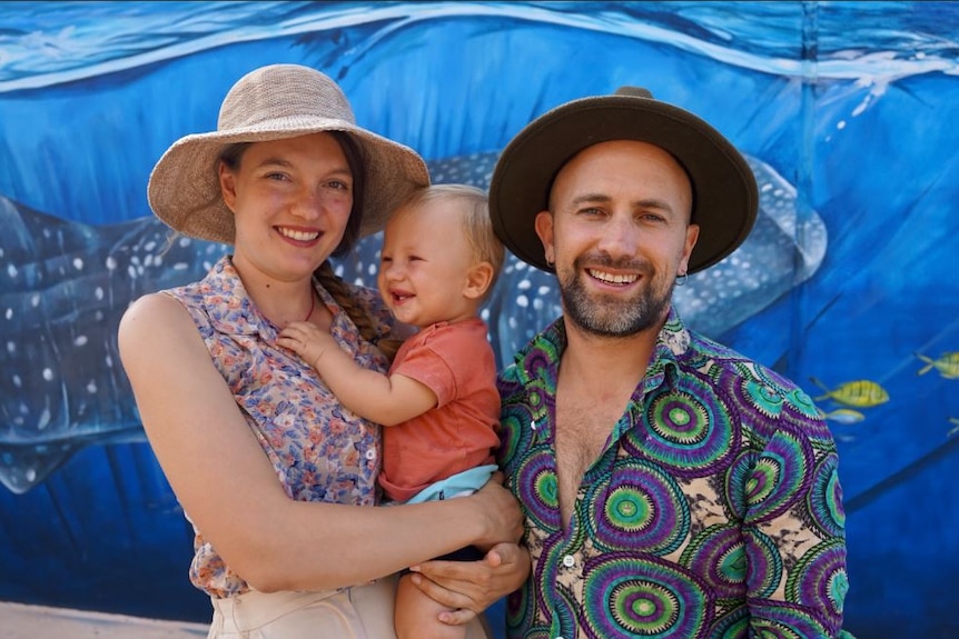 A man and woman wearing hats and colourful shirts holding a baby between them.