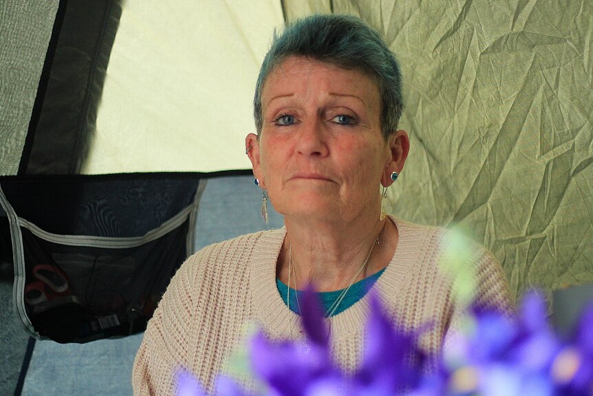 An older woman with blue hair, inside a tent. Flowers are in the foreground.