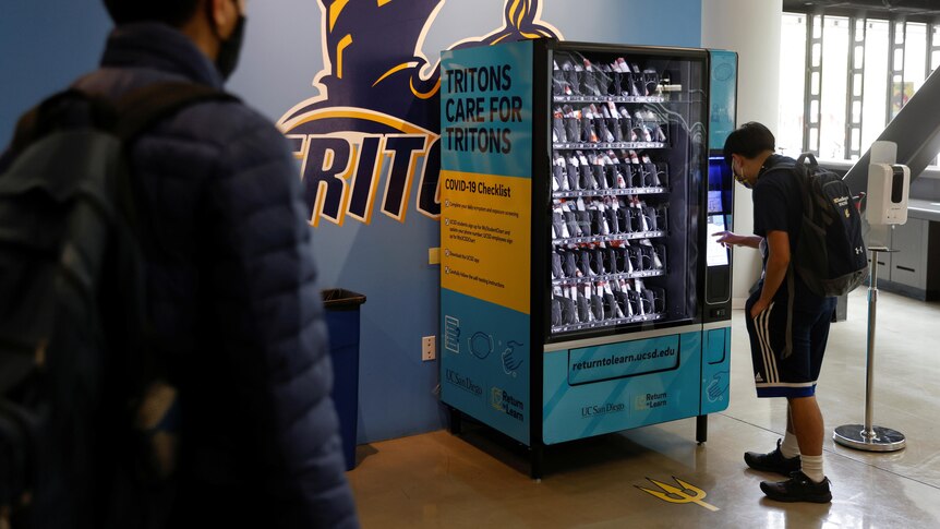 One man watches as another uses a vending machine that sells COVID tests. The sign says TRITONS CARE FOR TRITONS