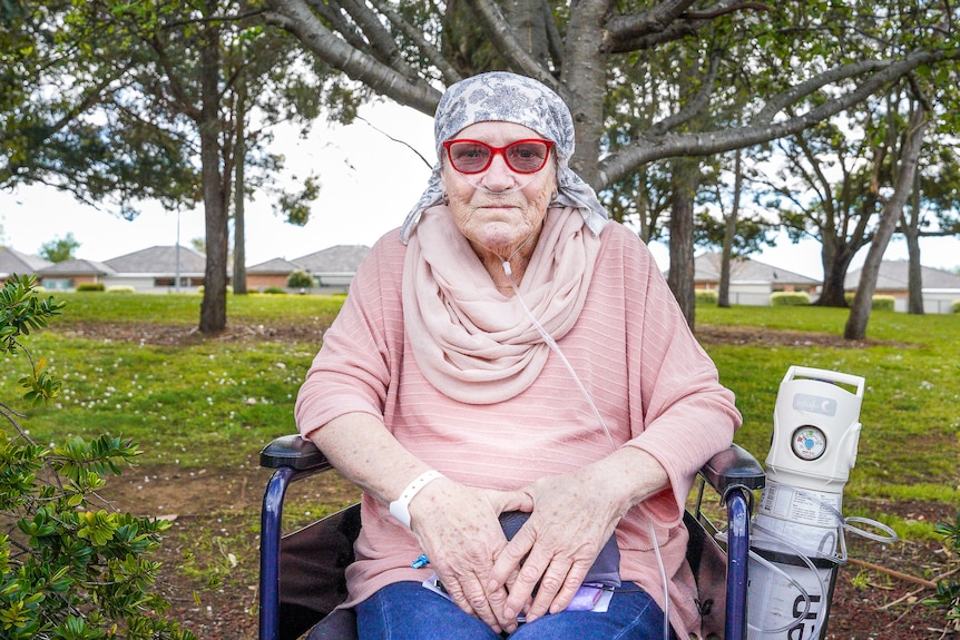 A woman with a shaved head wearing red sunglasses laughs in a wheelchair in a sunny courtyard.