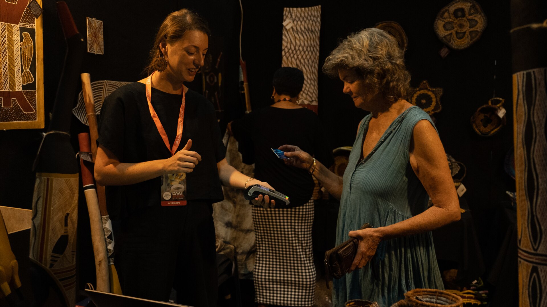 A woman taps her card on a card reader held by another woman, in front of black walls hung with Aboriginal art works.
