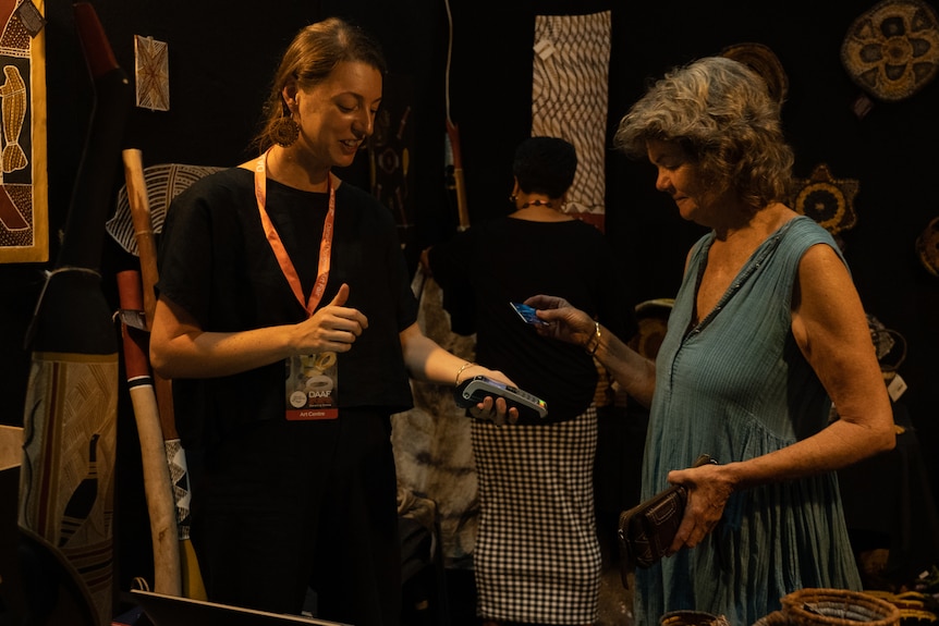 A woman taps her card on a card reader held by another woman, in front of black walls hung with Aboriginal art works.