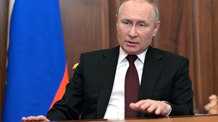 Vladimir Putin sits at a table wearing a maroon tie, white shirt and black suit.