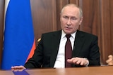 Vladimir Putin sits at a table wearing a maroon tie, white shirt and black suit.