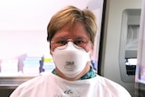 A woman with smiling eyes and short, fair hair in PPE and a face mask. She is wearing wire-rimmed glasses
