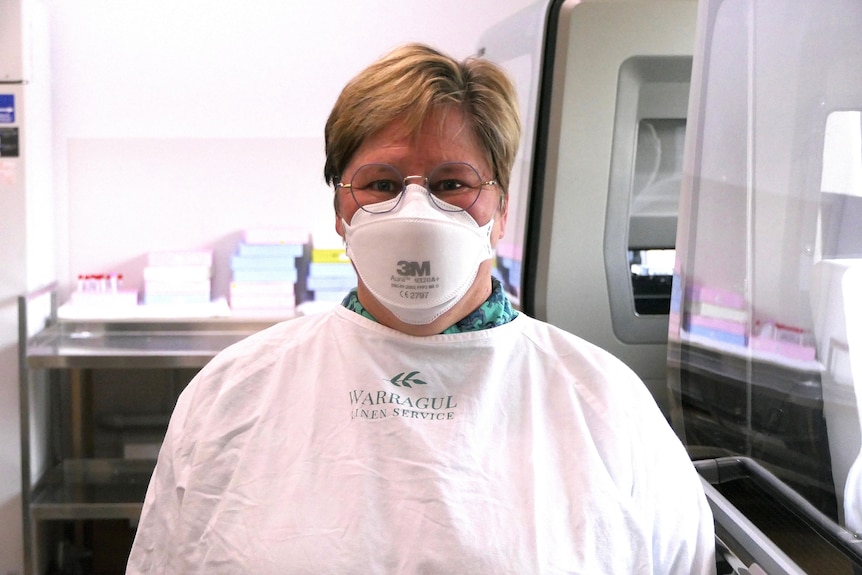 A woman with smiling eyes and short, fair hair in PPE and a face mask. She is wearing wire-rimmed glasses