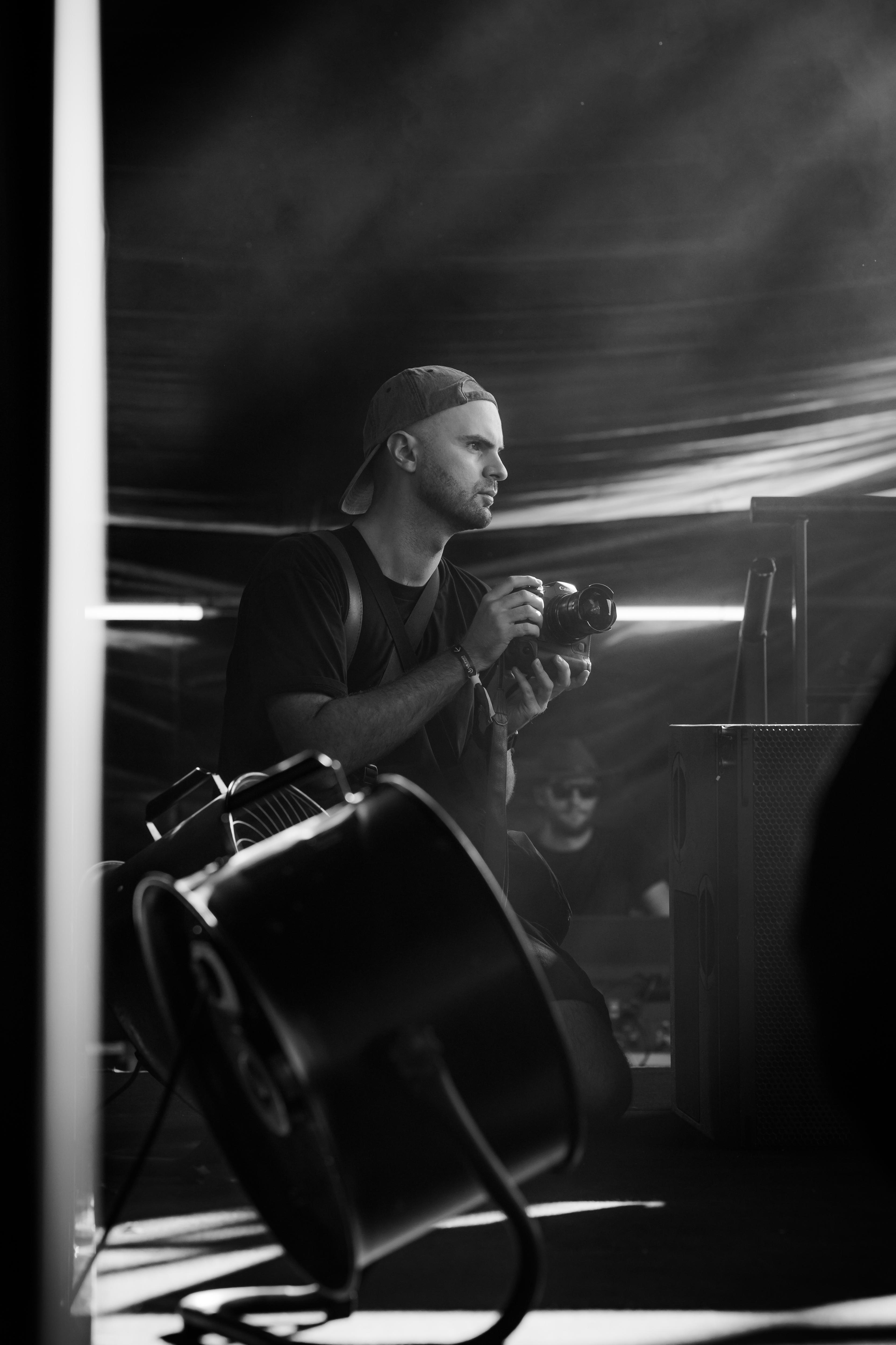 A black and white photo of a photographer with a cap on his hat backwards and holding a camera.