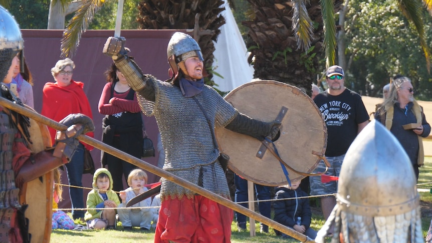 A man in chainmail armour holds up a sword and shield. A crowd of people are watching.