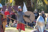 A man in chainmail armour holds up a sword and shield. A crowd of people are watching.