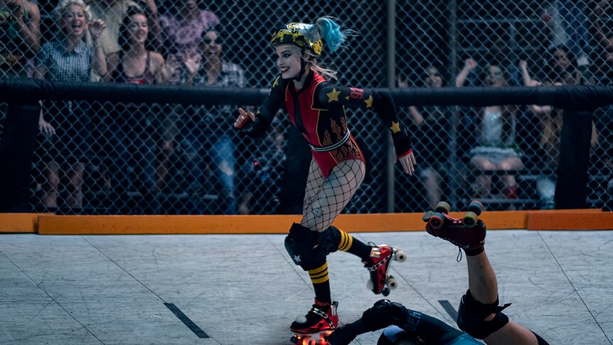 A woman with blonde and blue hair, skates past falling person in caged rink in long sleeve leotard and light up roller skates.
