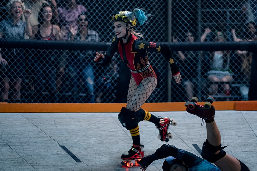 A woman with blonde and blue hair, skates past falling person in caged rink in long sleeve leotard and light up roller skates.