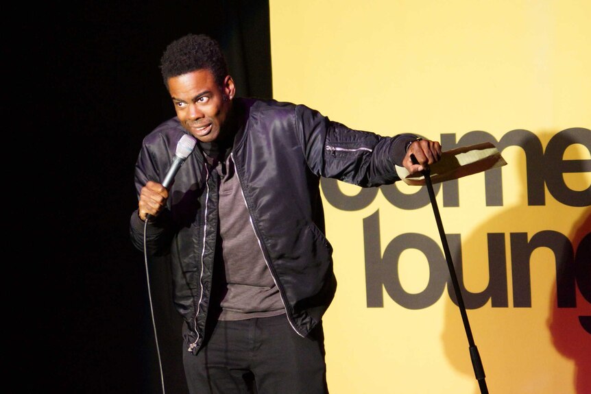American comedian Chris Rock talks into a microphone on stage at the Charles Hotel in North Perth with a yellow sign behind him.