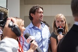 A man in a blue shirt is surrounded by reporters with microphones and cameras. 