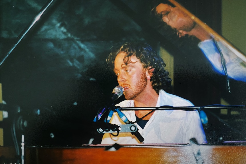 A young man with curly hair sings into a microphone while playing piano.