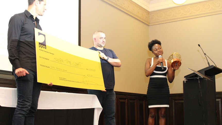 Sampa Tembo speaks into the microphone holding her trophy next to two men with a giant cheque
