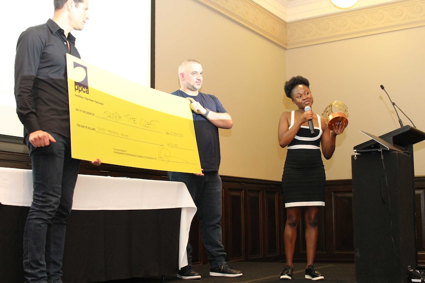 Sampa Tembo speaks into the microphone holding her trophy next to two men with a giant cheque