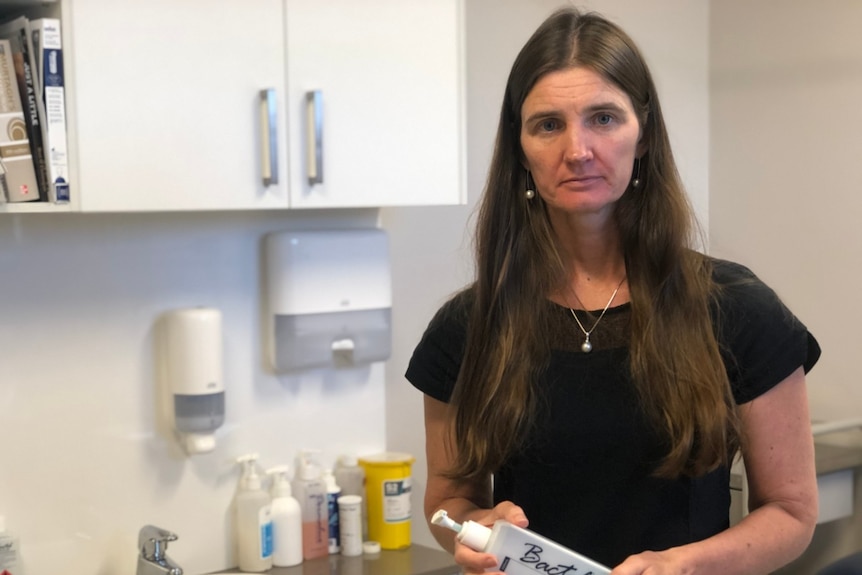 A middle-aged woman with long, brown hair, dressed in black, holds a bottle of hand sanitiser