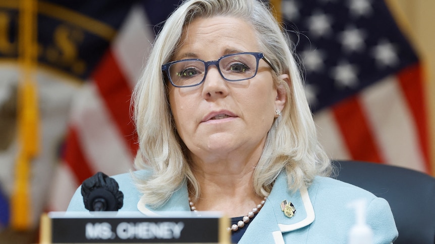 Liz Cheney, in a pale blue blazer with white trim, sits at a desk with a name plate, in front of a US flag