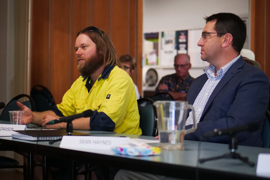 A man in a yellow vest sitting at a table next to a man in a suit