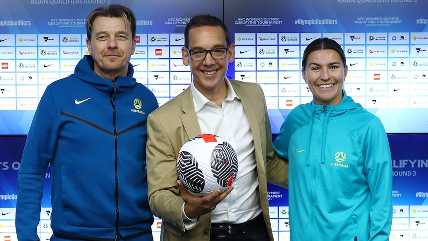 Steve Dimopoulos holding a soccer ball with Matildas head coach Tony Gustavsson and player Steph Catley.