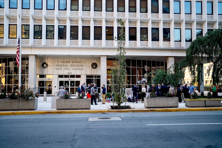 A line of people stands outside a large building with 'Caleb Boggs Federal Building' signed above glass doors.