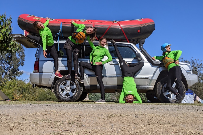 Four women with various fun poses standing in front of a four wheel drive, with a raft on top of the car.