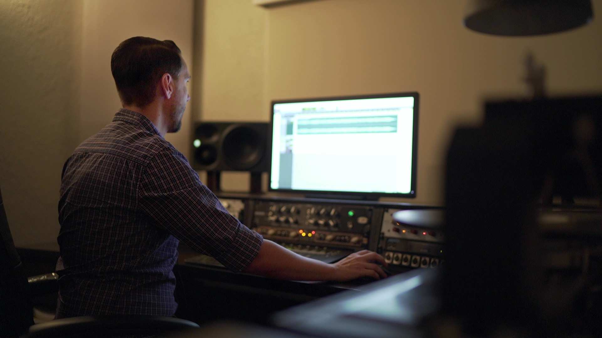 A man sits in a dimly lit room working on audio files on a computer