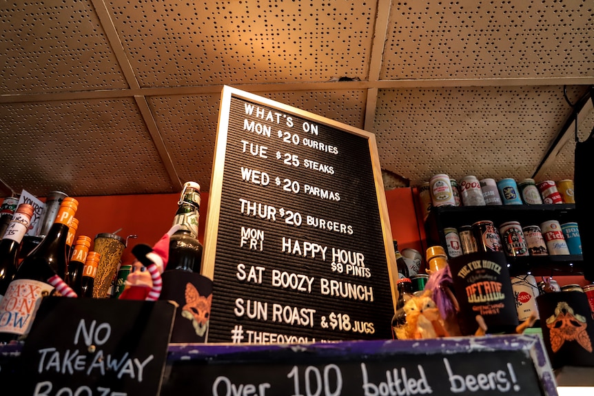 A black board with white text listing pub specials, sitting atop a fridge amid rows of beer cans on pub shelves   
