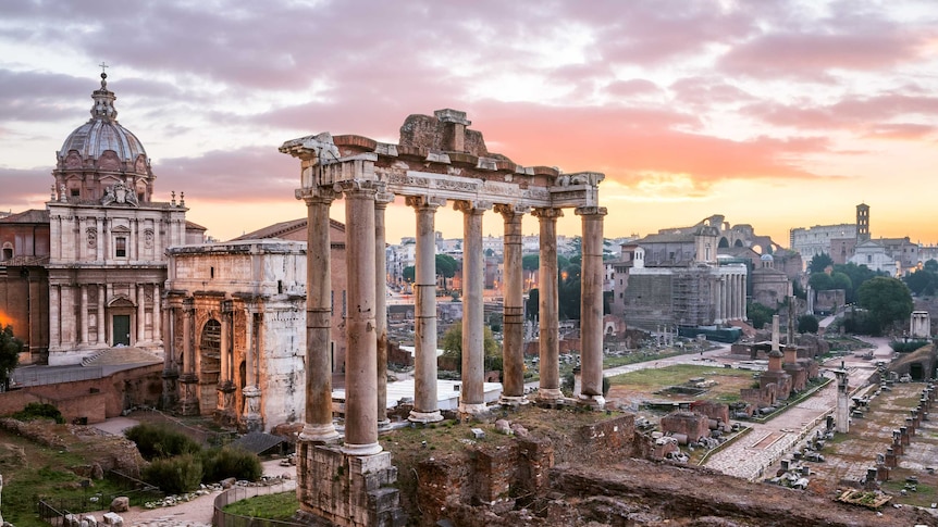 Orange and purple clouds behind ancient ruins, large columns, cobblestone paths and a large basilica.